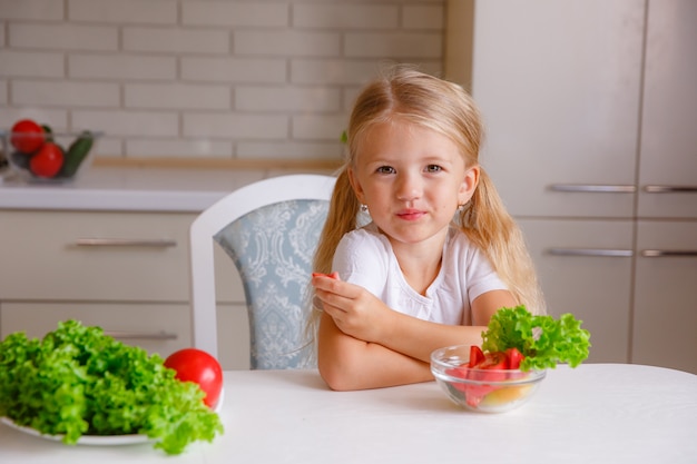 blonde kind in de hoed van een chef-kok in de keuken groenten eten