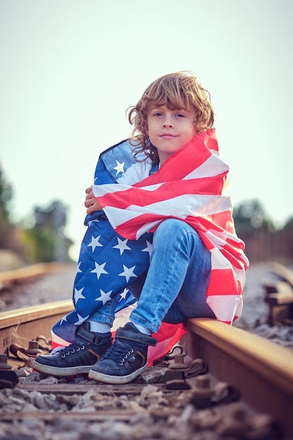 Blonde kind kijkt naar de camera terwijl hij op de spoorlijn zit tijdens de Amerikaanse Onafhankelijkheidsdag op een wazige achtergrond