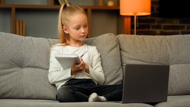 Blonde kleine slimme schoolmeisje leerling luistert naar educatieve les op laptop thuis online les