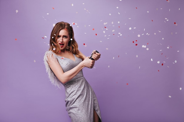 Blonde lady in silver dress throwing confetti portrait of curly\
attractive woman in festive outfit looking into camera and having\
fun on purple background