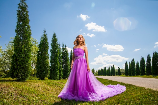 Blonde lady in violet dress with long skirt near row of green trees in sunlight