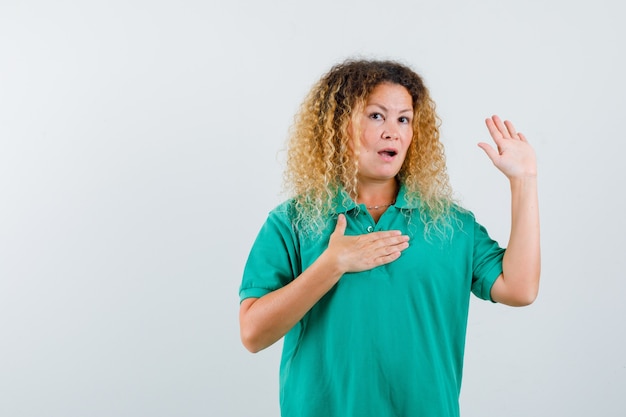 Blonde lady with curly hair showing stop gesture, keeping hand on chest in green T-shirt and looking puzzled. front view.