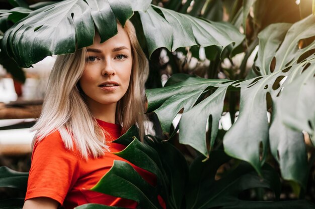 Blonde lady with natural makeup posing in rainforest. Gorgeous caucasian woman in red t shirt standing among monstera leaves, enjoying summer vacation. Wildlife and people concept.