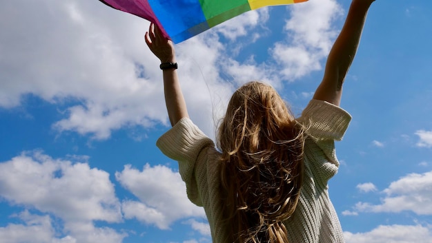 Blonde lesbian woman holding a rainbow lgbt gender identity flag on sky background with clouds on a