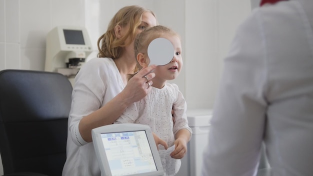 Blonde little girl closes one eye  Optometrist in clinic checking little childs vision