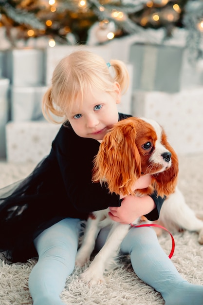 Blonde little girl hugs a dog on the background of a christmas tree and gifts