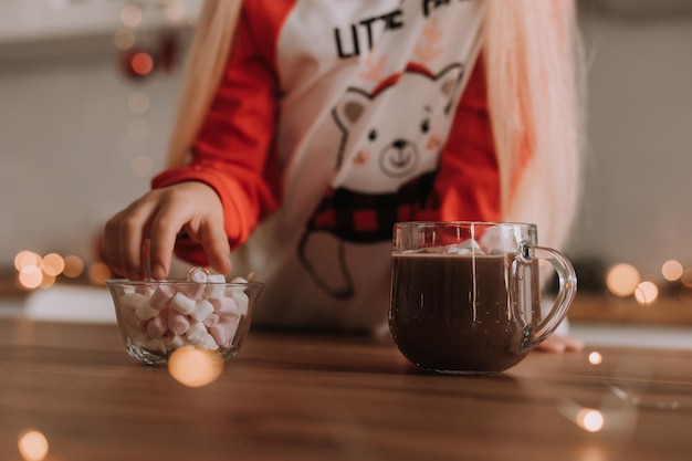 Blonde little girl in red Christmas pajamas adds marshmallows to a mug with a hot drink in a beautifully decorated kitchen for the holiday. lifestyle. space for text. High quality photo