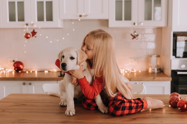 La bambina bionda in pigiama natalizio rosso è seduta in una cucina inghirlandata con un cane bianco. fine settimana e vacanze invernali in una calda cerchia familiare. bambino e animale domestico. spazio per il testo. foto di alta qualità