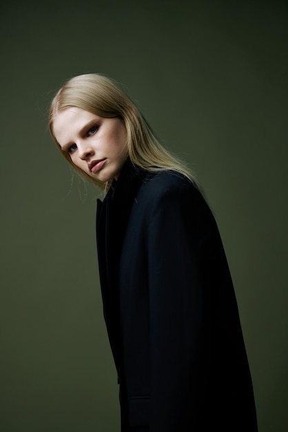 금발은 녹색 배경에 스튜디오에서 반으로 포즈를 취하는 검은 재킷을 입고 그녀의 어깨 너머로 보입니다. 세련된 패션 사진의 개념