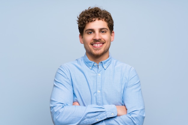 Photo blonde man over blue wall keeping the arms crossed in frontal position
