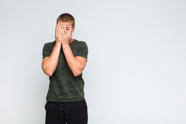 Blonde  man covering face with hands, peeking between fingers with surprised expression and looking to the side