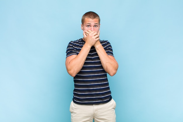 Blonde  man covering mouth with hands with a shocked, surprised expression, keeping a secret or saying oops