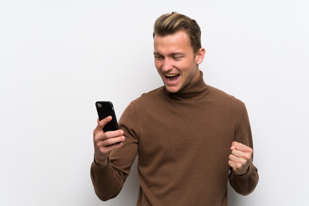 Blonde man isolated white wall celebrating a victory with a mobile