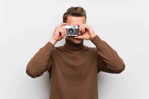 Blonde man over isolated white wall holding a camera