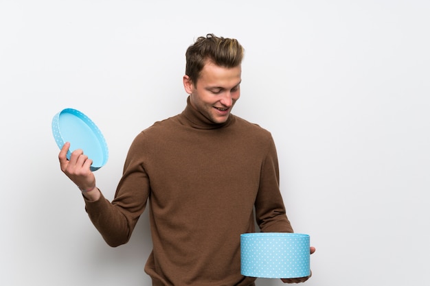 Blonde man over isolated white wall holding a gift in hands