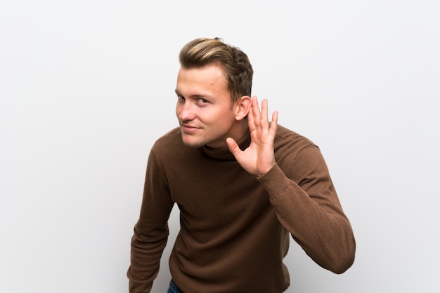 Uomo biondo sopra isolato muro bianco ascoltando qualcosa mettendo la mano sull'orecchio