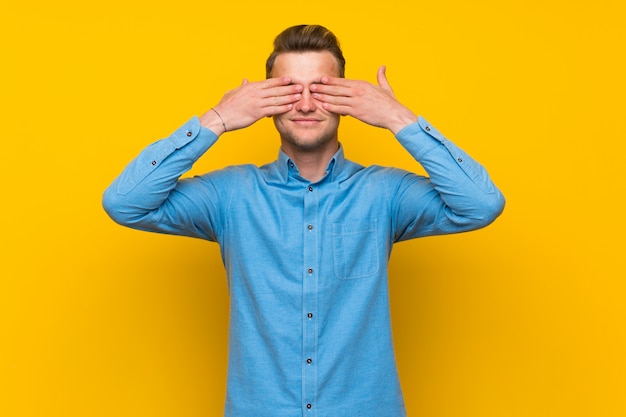 Blonde man over isolated yellow wall covering eyes by hands