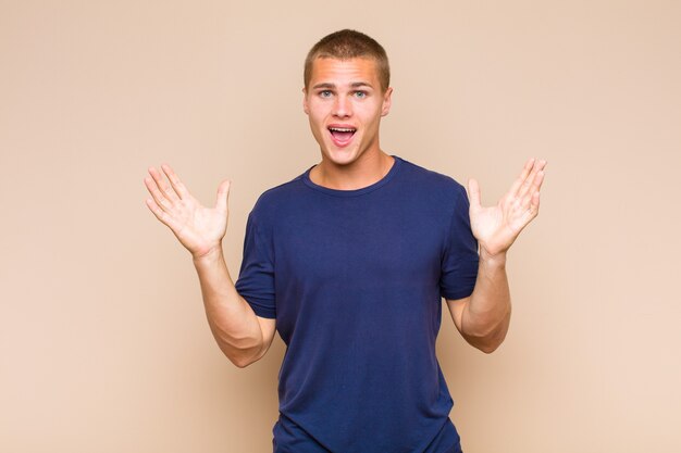 Blonde man looking happy and excited, shocked with an unexpected surprise with both hands open next to face