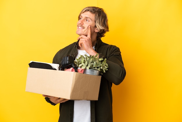 Blonde man making a move while picking up a box full of things isolated on yellow background thinking an idea while looking up