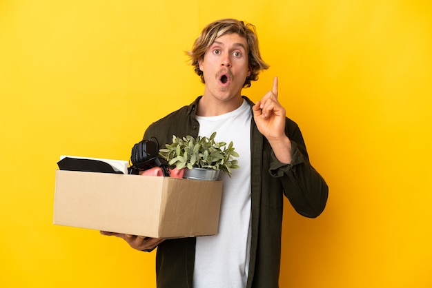 Blonde man making a move while picking up a box full of things isolated on yellow intending to realizes the solution while lifting a finger up