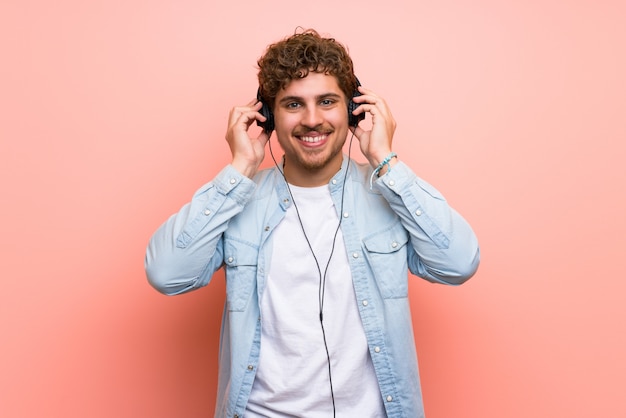 Blonde man over pink wall listening to music with headphones