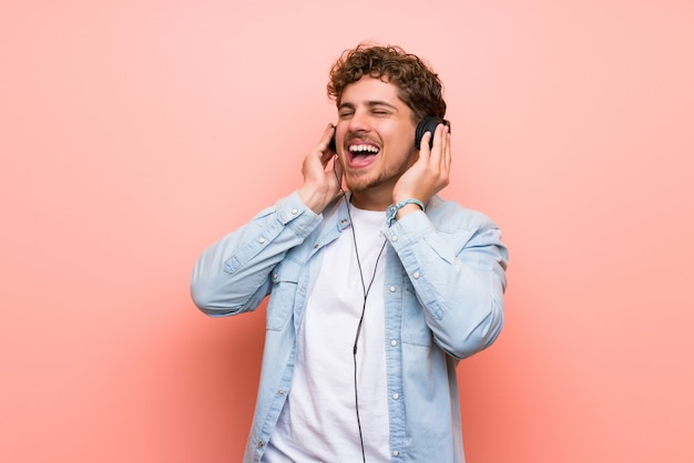 Blonde man over pink wall listening to music with headphones