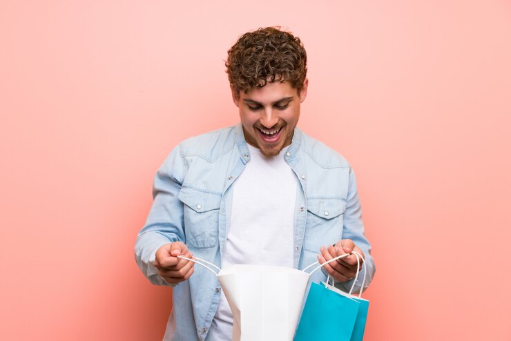 Photo blonde man over pink wall surprised while holding a lot of shopping bags