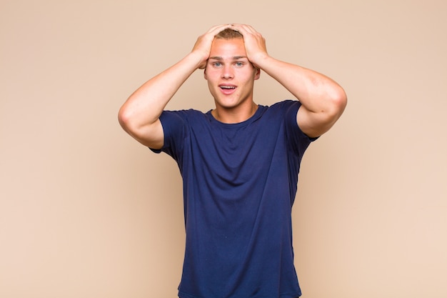 Blonde  man raising hands to head, open-mouthed, feeling extremely lucky, surprised, excited and happy