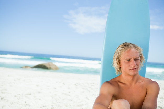 Blonde man sitting in front of his surfboard while looking towards the side