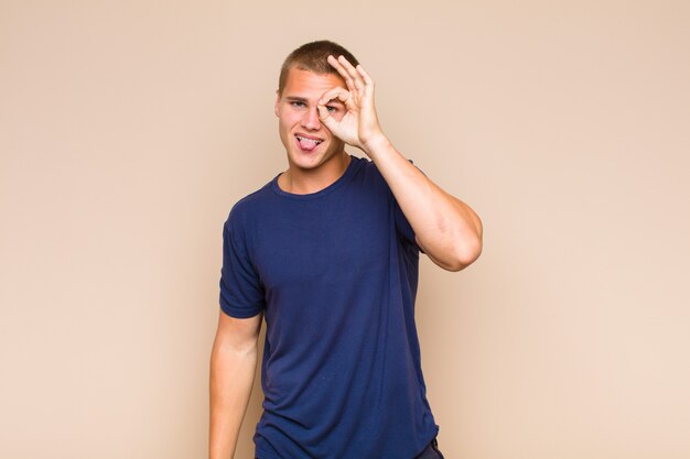 Blonde  man smiling happily with funny face, joking and looking through peephole, spying on secrets