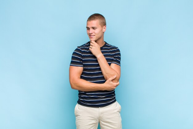 Blonde man smiling with a happy, confident expression with hand on chin, wondering and looking to the side