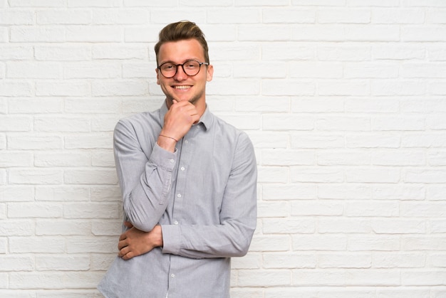 Blonde man over white brick wall with glasses and smiling