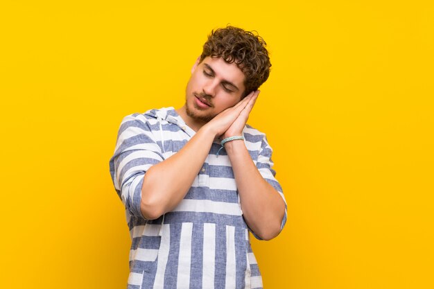 Uomo biondo sopra la parete gialla che fa gesto di sonno nell'espressione dorable