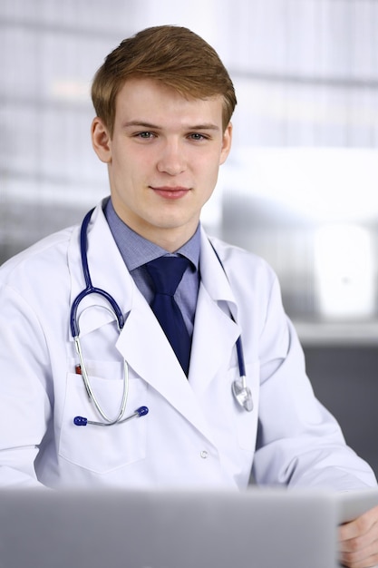 Blonde mannelijke arts zit en werkt met tabletcomputer in kliniek op zijn werkplek, close-up. Jonge arts aan het werk. Perfecte medische dienst, geneeskundeconcept.