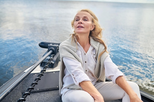 A blonde mature woman in white clothes sitting on a boat and looking dreamy