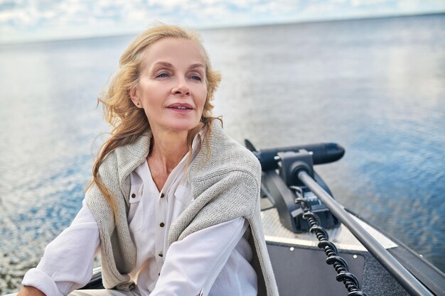 A blonde mature woman in white clothes sitting on a boat and looking dreamy