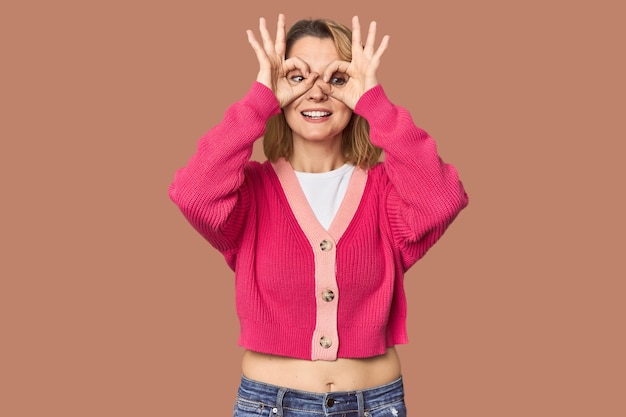 Blonde middleaged Caucasian woman in studio showing okay sign over eyes