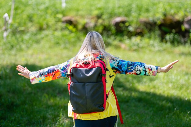 Blonde middleaged woman in bright clothes with a backpack in the park on a sunny day rear view