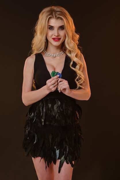 Блондинка-модель в черном платье с перьями и ожерельем. Она улыбается, показывает зеленые и голубые фишки, позирует на коричневом фоне. Покер, казино. Крупный план