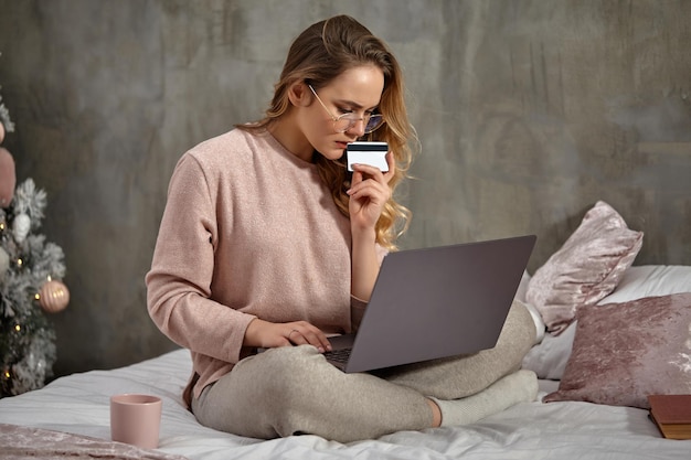 Блондинка-модель-блогер в очках в повседневной одежде держит пластиковую карту и работает на своем ноутбуке, сидя...