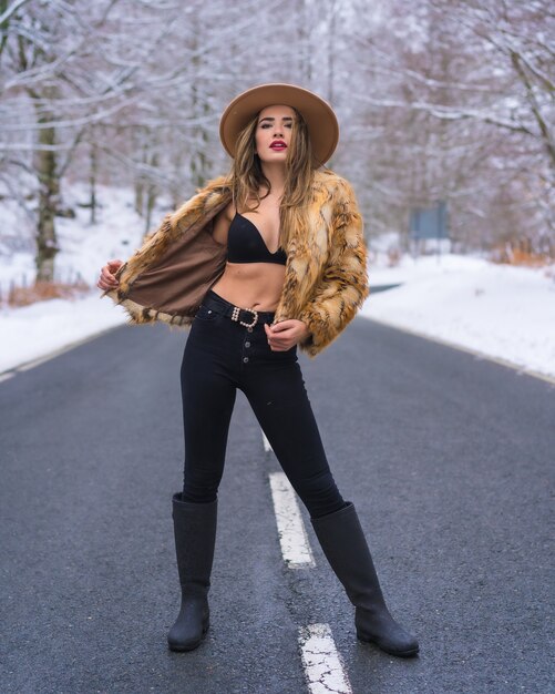 Ragazza bionda modello godendo in lingerie, un cappello da cowboy e un maglione leopardato in mezzo alla strada con alberi congelati sullo sfondo, stile di vita invernale