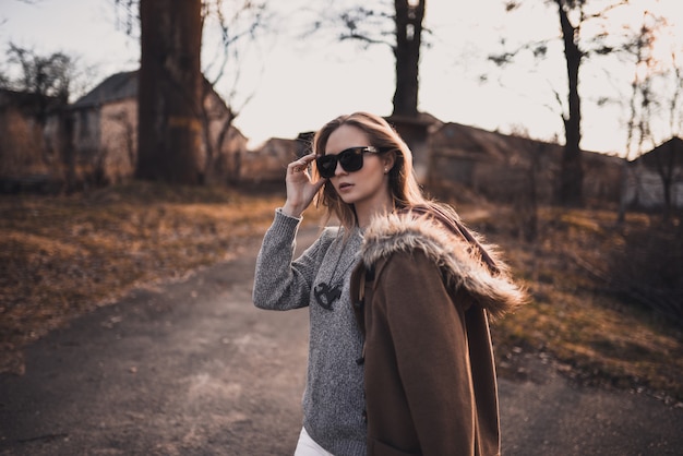 Ragazza bionda modello in pantaloni bianchi, occhiali da sole, cappotto marrone