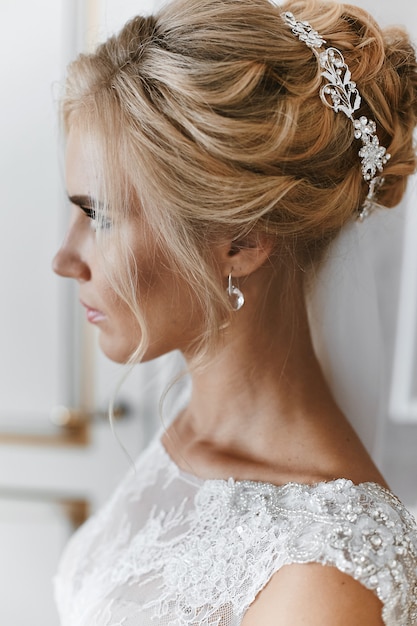 Blonde model girl with stylish wedding hairstyle and with jewelry in it, in lace white dress posing at interior, wedding preparation of the young bride