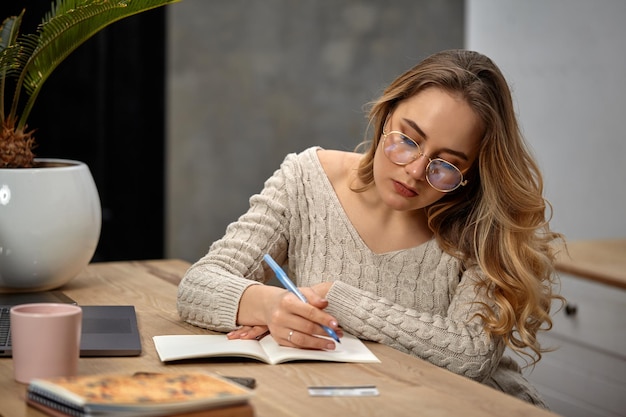 Blonde modelblogger in beige trui met bril zit in de keuken aan houten tafel en gaat wat schrijven...