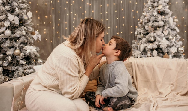 Blonde moeder zit op de bank en kust een jongetje op een achtergrond met een kerstboom