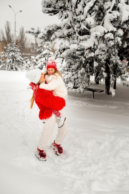 明るい服を着た金髪のお母さんは、冬の風景に対して娘を腕に抱きます。家族の表情。