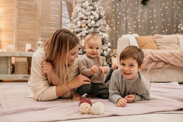 Blonde mom with young sons is sitting on a blanket near the Christmas tree in the room