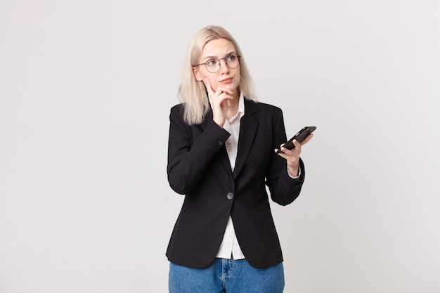 Blonde mooie vrouw denkt, voelt zich twijfelachtig en verward en houdt een mobiele telefoon vast