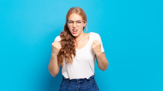 Blonde mooie vrouw die agressief schreeuwt met een geïrriteerde, gefrustreerde, boze blik en strakke vuisten, woedend voelend