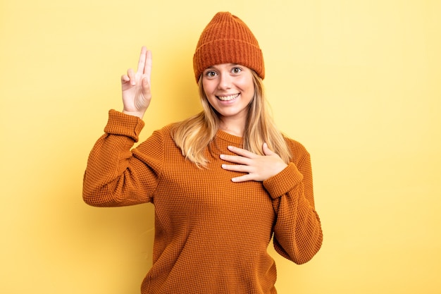 Blonde mooie vrouw die er gelukkig, zelfverzekerd en betrouwbaar uitziet, glimlacht en een overwinningsteken toont, met een positieve houding